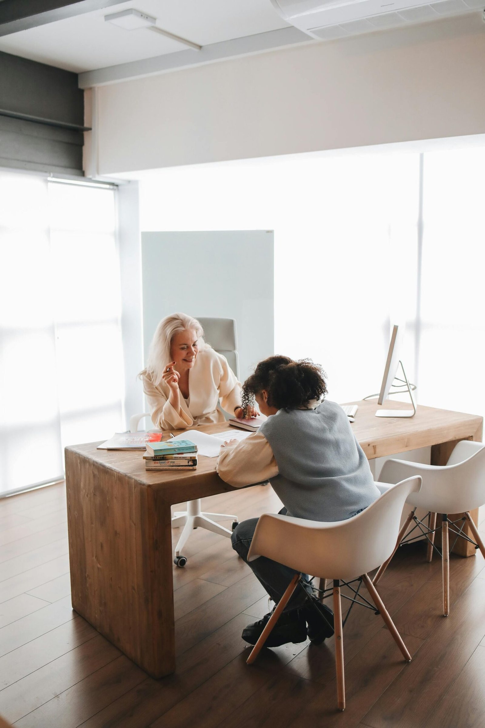 Home Two women engaged in a learning session at a wooden desk in a modern, bright office setting.